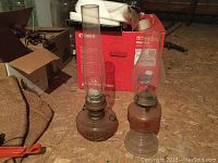 Photo of two antique oil lamps with tall glass chimneys, placed on a plywood surface surrounded by cardboard boxes and other items.