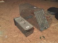 Photo showing two live animal traps placed on the ground; one large wire mesh trap with rear door, and one smaller trap with metal solid top and handle.