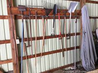 Wide shot of ten assorted garden tools hanging on wooden wall rack, displaying various types including rakes, shovels, hoes, and forks mostly with wooden handles and metal heads.