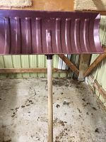 Purple large snow shovel with wooden handle standing vertically on a concrete floor against a metal wall.