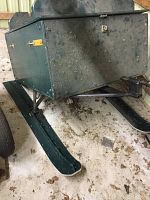 Front side view of vintage green metal snowmobile box sleigh on two skis with signs of wear and dirt on floor.
