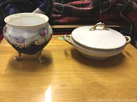Two porcelain items on a wooden surface; footed bowl with floral decoration and lidded serving dish with gold handles.
