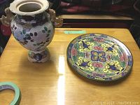 Photo showing a floral decorative vase with dual handles and a colorful decorative plate with intricate designs.