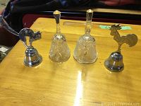 Four decorative dinner bells placed on a wooden surface, two metal animal-themed and two glass-cut style bells.