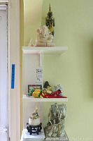 Three white wall-mounted shelves displaying assorted decorative items. Top shelf has a white elephant sculpture and a seated Buddha figurine. Middle shelf holds small Buddha statues, decorative eggs, and small figurines. Bottom shelf displays an Asian style sculpture of three horses.
