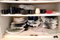 Two shelves of black bowls, cups, white cream and sugar pots, ashtrays, and plates in a cabinet.