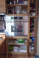 Kitchen cabinet section with stainless steel oven and wooden shelves containing plastic containers, pitchers, and thermos flask.