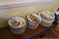 Three stacks of Sigma collectible plates showing assorted designs including bird art, floral patterns, and painted human & dog scene.