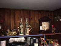 Wide view showing two ceramic beer steins, two figurines of women, brass eagle, brass dog, decorative pottery under warm lighting on wooden shelves.