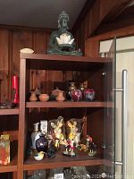 Photo showing large Buddha statue on top shelf holding lotus flower candle holder plus pottery jars, brass vases, and figurines on shelves below.