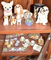 Photo showing 5 canine figurines on a wooden shelf including a stuffed white dog with a checkered scarf and 4 ceramic or resin dog figurines of different breeds. Also visible are two glass paperweights in front of the dogs and assorted miniature decorative items in a cabinet below.