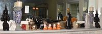 Wide view of mantle shelf showing full lot including candle stands, black Thai-themed figurines, silver animal figures, orange figurines, and decorative bottles.