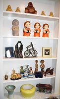 Five shelves containing various figurines including Buddha statues, three orange-robed monk figurines, abstract human sculptures, a bicycle figure, small decorative items, pottery vases.