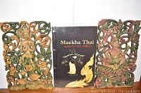 Two carved wooden Thai wall art pieces with a Maekha Thai menu positioned between them on a wooden surface.