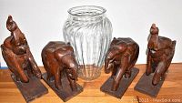 Four carved wooden elephants arranged around a tall ribbed glass vase on white ground