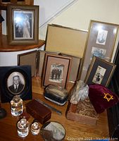 Group of vintage framed photographs, glass inkwells, wooden box, brush, and other small memorabilia arranged on a floor under stairs.