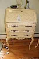 Wood secretary desk with drop front and three drawers, ornate metal handles, curved cabriole legs, painted cream with decorative accents.