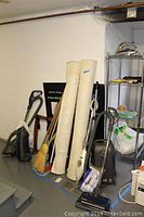 Photo showing Kenmore canister vacuum, Shark wand vacuum, broom, Swiffer mop, two rolled area rugs, and shelving with cleaning supplies in a basement setting.