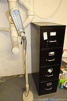 Photo of a black metal filing cabinet with four drawers next to a tall adjustable arm lamp in a basement setting.