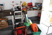 Wide shot of garage corner showing hand truck, shop vacuum, plastic storage bins, toolboxes and ladder against workbench with cords and tools.