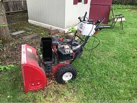 Side view of the red metal auger housing, black engine, and white wheel of the snow blower showing signs of wear and rust on grassy lawn.