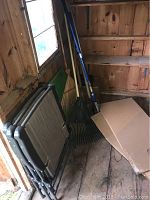 Photo showing 3 black metal base folding lawn chairs leaning against a wall, with garden rakes alongside and cardboard on floor.