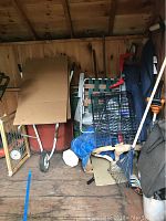 Corner of shed showing red metal wheelbarrow, wooden baby gate, two white metal folding chairs, blue rake, black container, and other garden tools stacked and leaning.