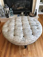 Front view of round tufted ottoman, showing script printed upholstery and tufted details with button accents, on hardwood floor in front of fireplace.