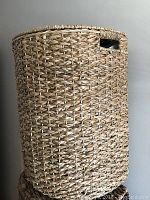 Single tall woven laundry hamper in light brown natural fibers with lid and side handle cutouts.