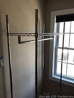 Side angled view showing stainless steel frame, wire shelves, and horizontal hanging bars by a window.