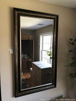 Vertical rectangular mirror hanging on a beige wall showing reflection of a room with window and furniture.