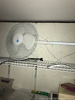 White pedestal fan placed on wire shelf, showing adjustable height and power cord