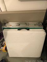 Three folding tables folded and stacked: one with white plastic top, one with green playing field top, and one aluminum framed 8 ft table.