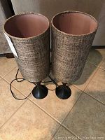 Two tall table lamps with black bases and woven textured cylindrical shades standing on tiled floor.