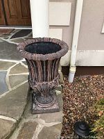 Front angle of one black and terra cotta cast plastic outdoor urn on stone and gravel surface near column and wall.