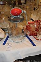 Full view of the vintage Dazey butter churn with glass jar and red crank handle on top, placed among other glassware.