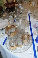 Photo of vintage glassware lot including gold trimmed pitcher and glasses, sugar, creamer, serving tray, martini mixer, wine decanter, and goblets arranged on table.