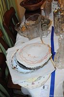 Photo of stacked vintage porcelain serving dishes, some with floral patterns and scalloped edges, placed on a white cloth on a table.