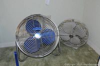Pair of fans on carpeted floor against wall; blue Patton fan with metal cage on left, off-white vintage GE fan on right.