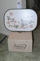 Photo of floral lap tray showing flower design and green trim, placed on top of original box labeled Anchor Hocking.