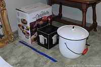 Columbian enamel chamber pot with lid, black rim, red handle, food dehydrator and hot pot in original boxes on green carpet near table and mirror.