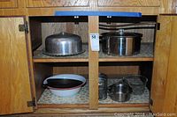 Closed cabinet with miscellaneous kitchen items such as metal cake dome, cooking pot, enamel bowl, cast iron skillet, wire baskets, and glass jars.