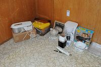 Photo showing all items arranged on a linoleum floor against a wood-paneled wall: White Magic ice cream freezer, yogurt maker, electric juicer, electric knife, Mr. Coffee maker, electric hand mixer, plastic container, and a cookbook on a stand.