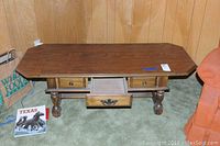 Full view of the coffee table with drop leaves extended and drawer open, placed against wood panel walls on carpeted floor.