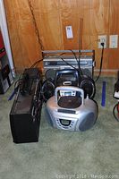 Photo of multiple radios including a black and silver GE stereo boom box, several portable CD players, a Panasonic radio, and power supply unit.