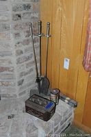 Wide shot showing fireplace tool set with shovel, sweeper, poker next to vintage cigar box filled with match boxes, also cylindrical can and fireplace broom.