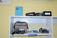 Shelf showing multiple cameras with cases and bags, Pana-Vue slide viewer box on top shelf, and black box labeled 'Converters' with electrical adapters.