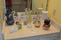 Various decorative items on white countertop including glass vases, porcelain figurines, ceramic urn, wooden box, and ceramic hen figurine.