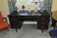 Front view of dark wood desk with six drawers and two slide-out surfaces. Several table lamps placed on top.