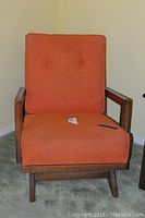 Front view of rocking armchair with orange cushioned seat and back, wooden arms and legs.